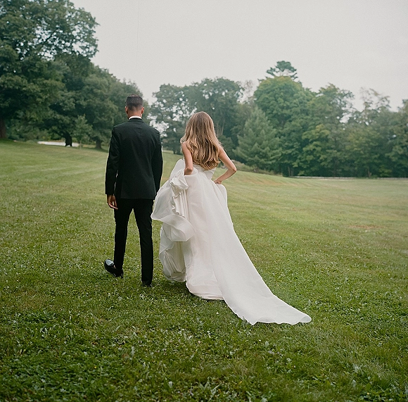 Couple portrait of bride and groom walking away, bride holding her wedding dress train across a grassy field under overcast sky