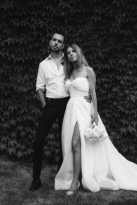 Couple portrait in a black and white wedding portrait, bride in strapless slit gown holding a rose bouquet beside groom by an ivy wall
