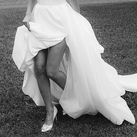 Wedding dress close-up showing a high slit wedding dress with strapless satin skirt, train, and pointed-toe pumps with jeweled buckle on grass lawn