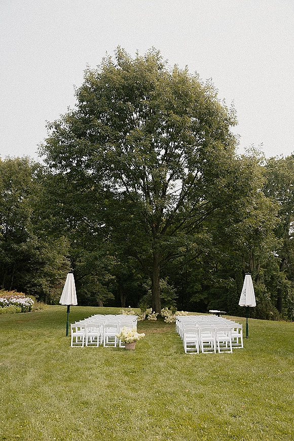 Outdoor ceremony setup with white folding chairs in rows and green floral arrangements on a grass lawn beneath a large tree, overcast sky