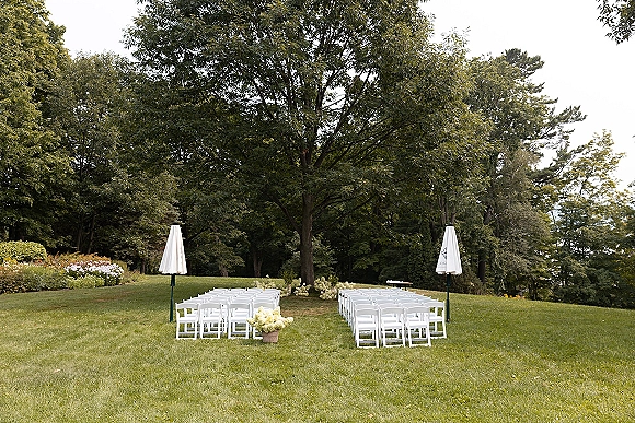 Ceremony setup with white folding chairs and floral arrangements lining a grass aisle beneath a large tree, with patio umbrellas nearby