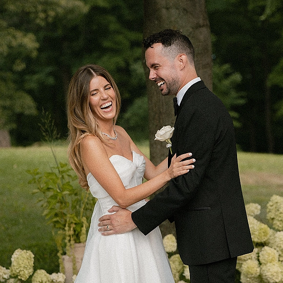 Couple portrait of bride and groom laughing in a garden, her strapless dress and necklace beside his tuxedo with white rose boutonniere