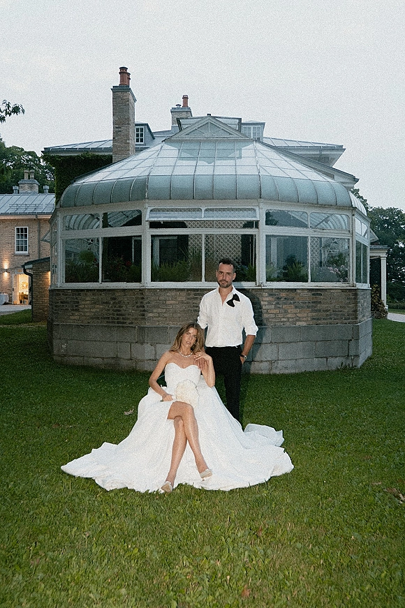 Couple portrait with bride sitting on grass in a strapless wedding dress as groom stands behind, conservatory and brick estate backdrop
