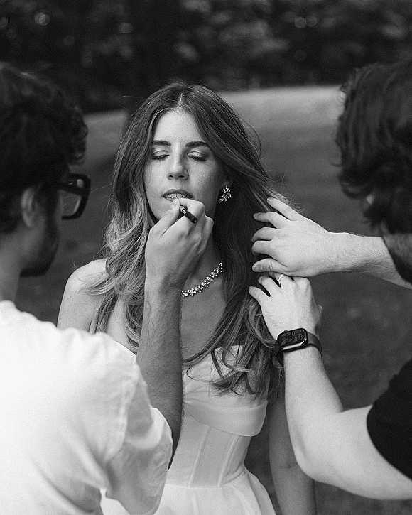 Bridal getting ready as bride receives makeup touch up, applying lipstick in a strapless dress with crystal earrings and necklace, greenery behind