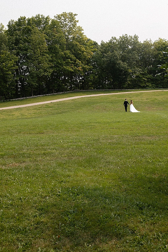 Couple portrait of bride and groom walking away hand in hand, her wedding dress train trailing on a curved path in a grassy field
