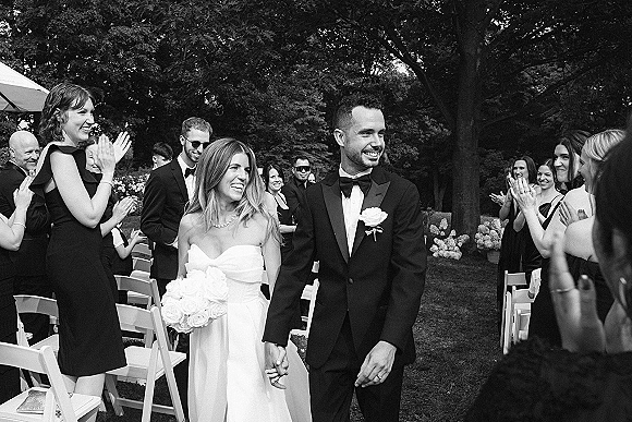 Wedding recessional as bride and groom walk the aisle holding hands, bride with rose bouquet, guests applauding in outdoor garden setting