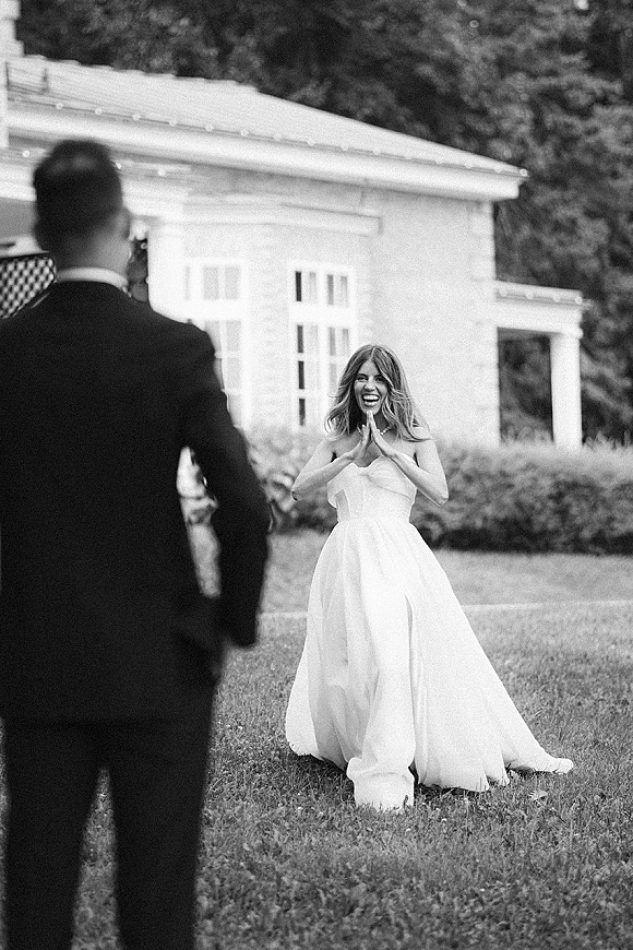 First look moment as the bride in a strapless gown and pearl necklace laughs toward the groom in a tuxedo on a lawn by a white building