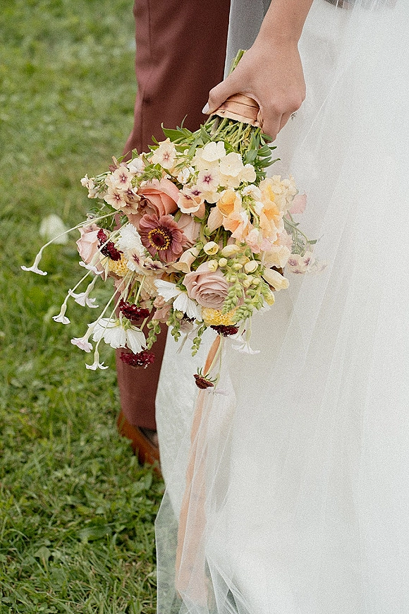 Bridal bouquet with a wildflower wedding bouquet mix of pastel blooms and greenery, wrapped in peach ribbon, held on a grassy lawn