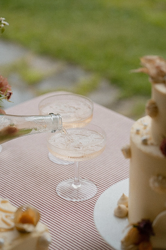 Champagne pour into a coupe glasses champagne tower beside mini cakes on a striped tablecloth, with flowers and greenery along a stone path