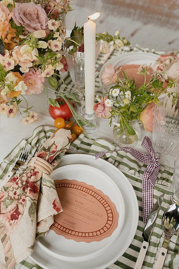 Reception tablescape with wedding place setting on a striped tablecloth, pastel floral centerpiece and taper candle among glassware and tomatoes