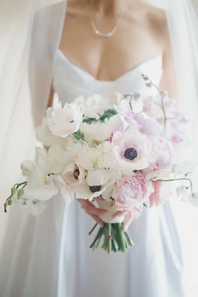 Bridal bouquet with white orchid blooms and blush peonies, held by a bride in a strapless dress and veil against soft neutral light backdrop