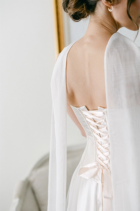 Wedding dress back with a corset back wedding dress laced in satin ribbon, cape sleeves framing her updo and pearl hoop earring by a mirror edge