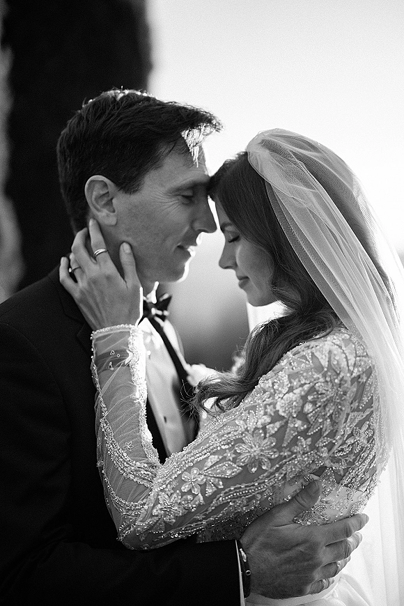 Wedding couple portrait in black and white with bride and groom embracing, foreheads touching, her veil and beaded lace gown against bright sky