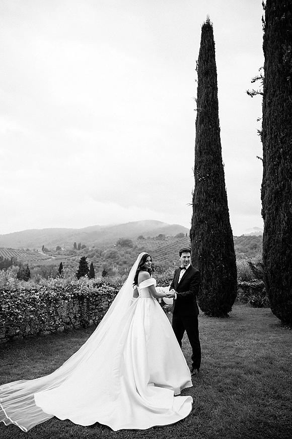 Couple portrait in a black and white wedding portrait, bride and groom holding hands on a lawn by cypress trees and hills, veil trailing