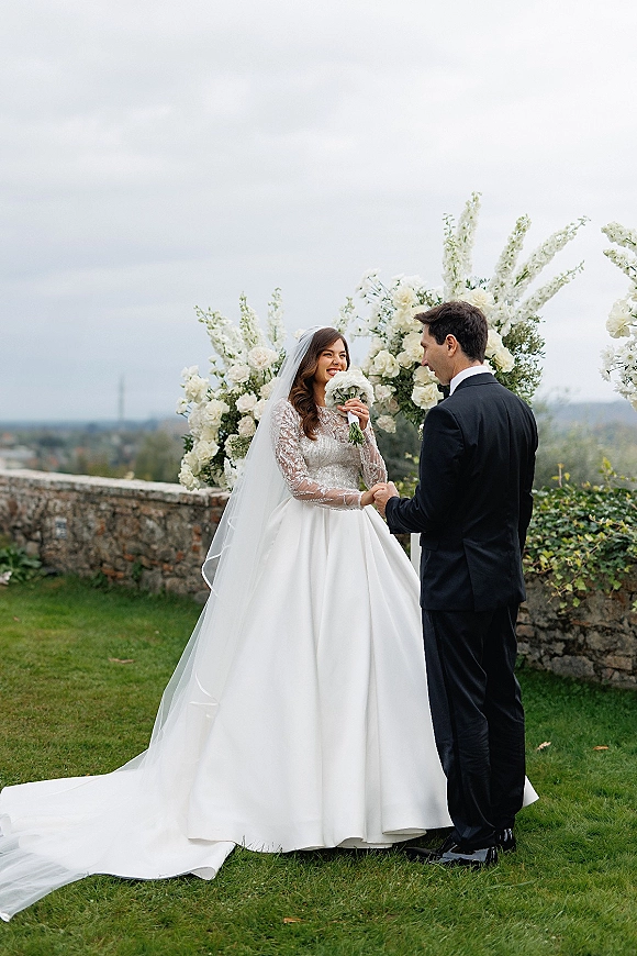 Couple portrait of bride and groom holding hands as she smells a white rose bouquet under a floral arch on a lawn by a stone wall