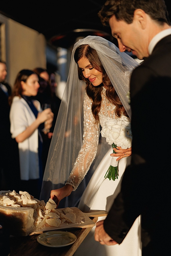 Wedding cake cutting as bride in long sleeve beaded dress and veil cuts a white cake with groom in tuxedo at an indoor reception