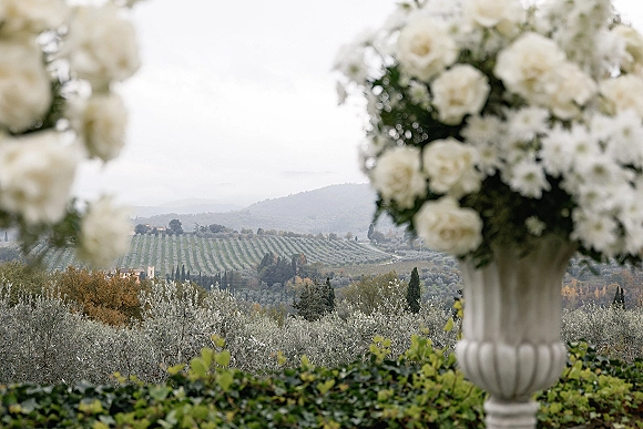 Wedding floral urns with white rose arrangements and greenery frame a ceremony entrance, set against cloudy vineyard hills and trees