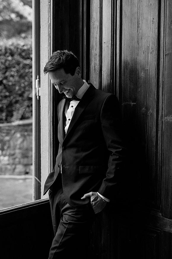 Groom portrait in tuxedo with black bow tie and pocket square, leaning by a window against a wood-paneled wall with greenery outside