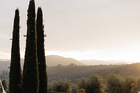 Wedding venue landscape with cypress trees framing rolling hills and a valley, lit by warm sunset light under an open sky