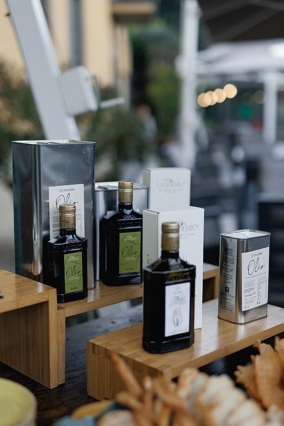 Wedding favors of olive oil bottles in metal tins with branded labels, styled on wooden risers on an outdoor patio with string lights