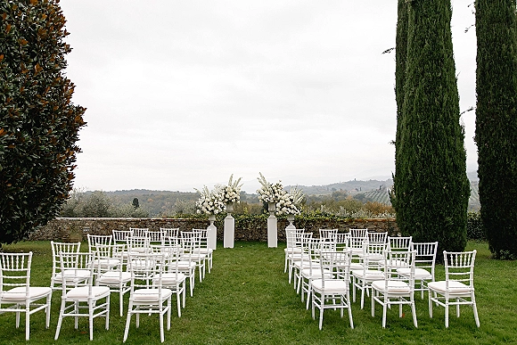 Outdoor ceremony setup with garden wedding ceremony white chiavari chairs lining an aisle, floral pedestals by a stone wall on a lawn