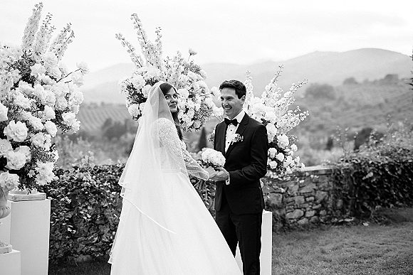 Ceremony moment as bride and groom hold hands under a white floral wedding arch, bouquet and veil flowing with mountains and vineyard hills behind