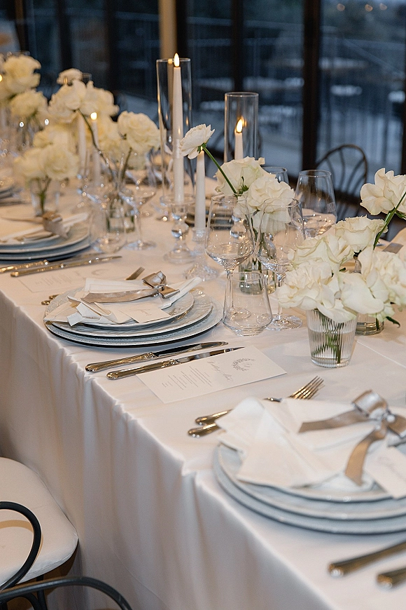 Reception tablescape with a white rose centerpiece, taper candles in glass hurricanes, layered plates and menus on a long table by windows