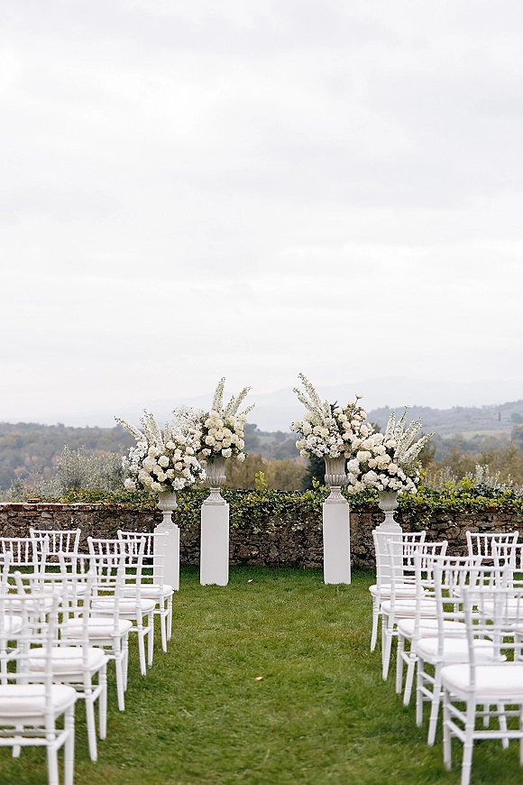 Ceremony setup with white floral arrangements lining a white chiavari chair aisle on a lawn, framed by stone wall and rolling hills