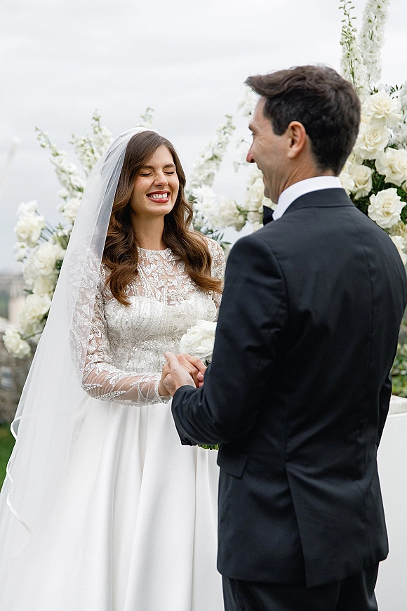 Wedding vows as bride in lace gown and groom in suit hold hands and smile beneath a floral arch with white roses outdoors