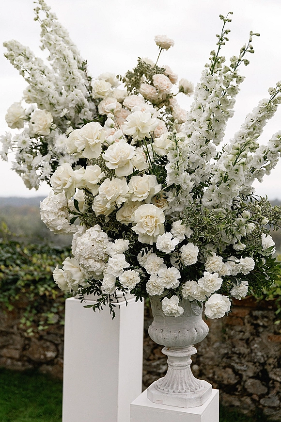 Wedding floral arrangement of white ceremony flowers in a stone urn on a pedestal, with greenery against a stone wall and hills beyond