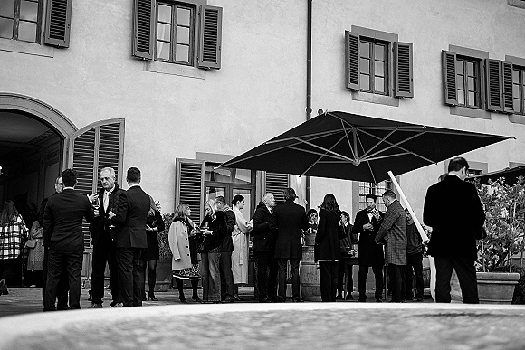 Wedding cocktail hour with wedding guests mingling in formal suits, holding champagne and wine beside a patio umbrella and barrel table in a villa courtyard