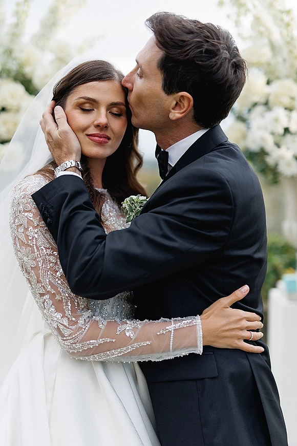 Wedding couple portrait of groom kissing bride’s forehead as they embrace, her veil and beaded long sleeves against greenery and white florals