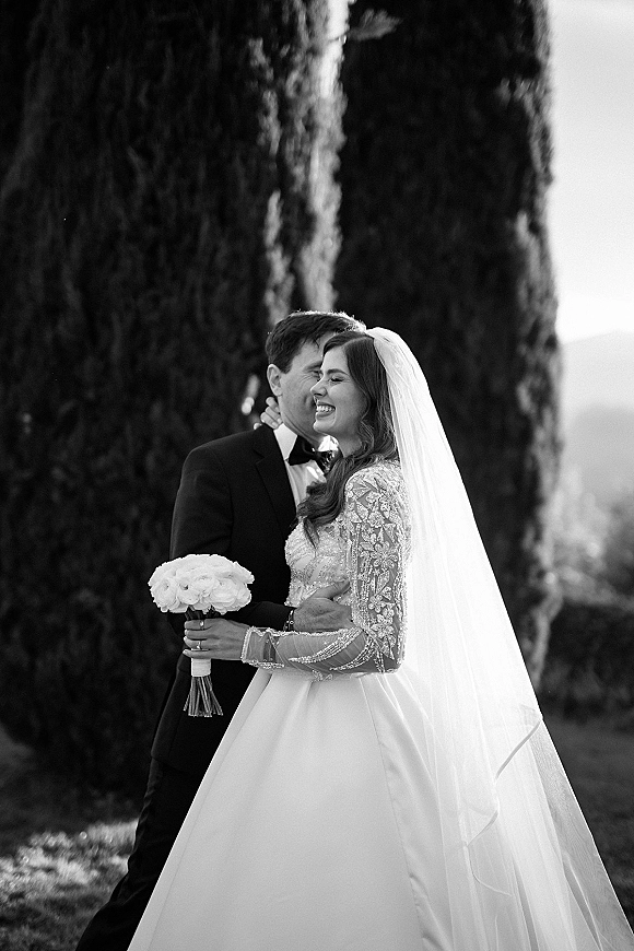 Couple portrait in a black and white wedding portrait as the groom hugs the smiling bride in a lace long sleeve dress with cathedral veil outdoors