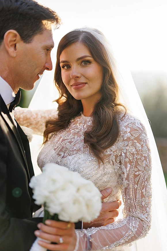 Wedding couple portrait of groom holding bride close as she looks at the camera, cathedral veil and white bouquet glowing in sunset light
