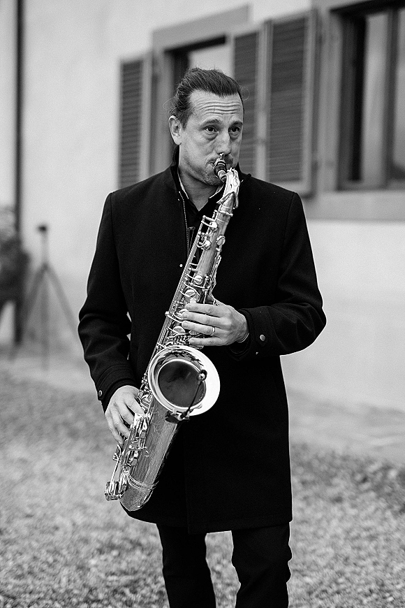 Wedding saxophonist portrait of a musician in a black coat holding a saxophone in a courtyard beside a building with shuttered windows