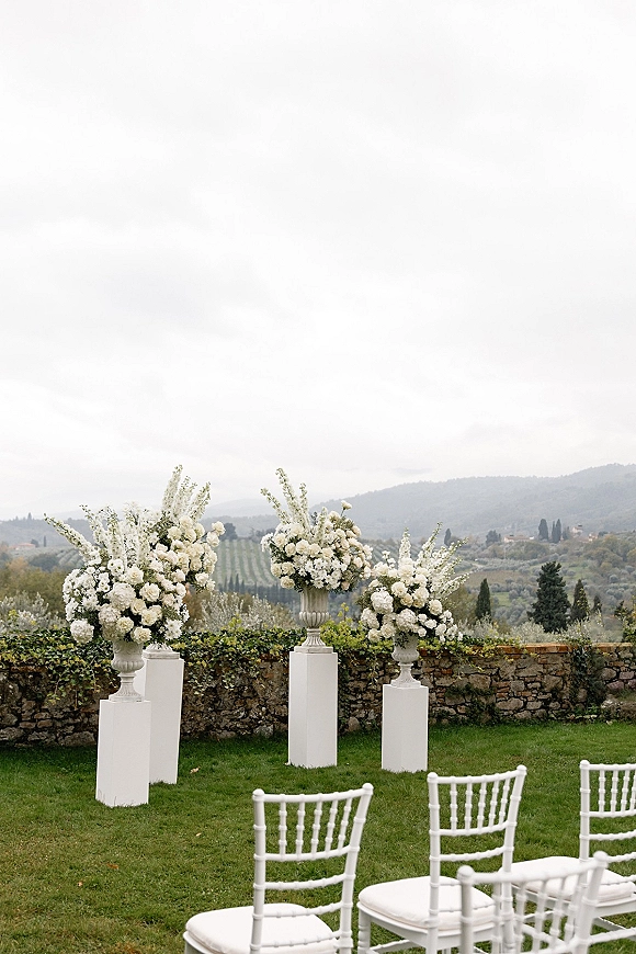 Ceremony floral arrangements with wedding aisle urns of white blooms on stone pedestals, flanking white Chiavari chairs on a lawn by a stone wall