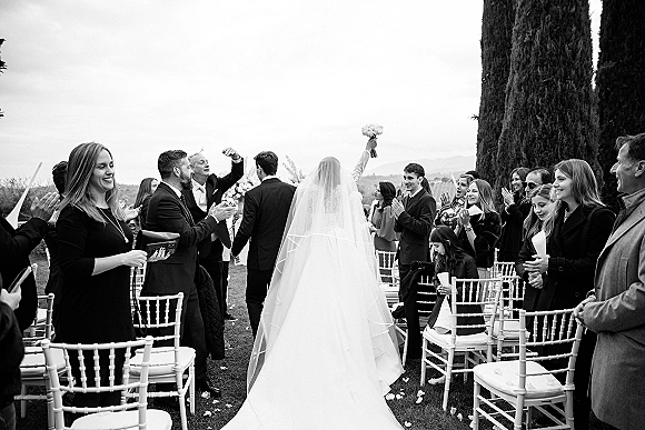 Wedding recessional as bride and groom walk down aisle, bride raising bouquet, long veil trailing, guests cheer among chairs on a tree-lined lawn