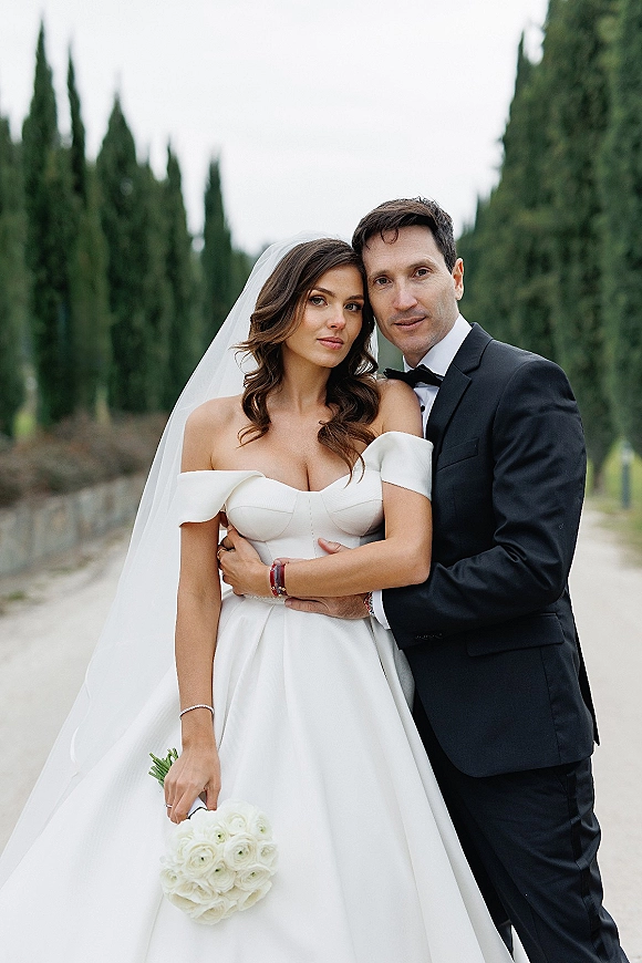 Couple portrait of bride and groom holding a bouquet, her long veil trailing on a tree-lined path with tall evergreens behind them