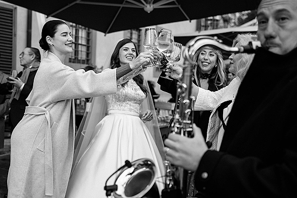 Wedding toast with champagne flute as bride in veil and friends clink glasses on an outdoor patio under umbrella canopy with guests behind