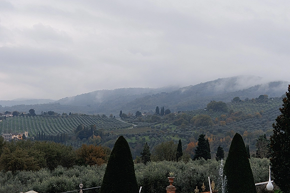 Wedding landscape with vineyard hills, olive and cypress trees, and distant buildings under a cloudy sky with misty mountains beyond