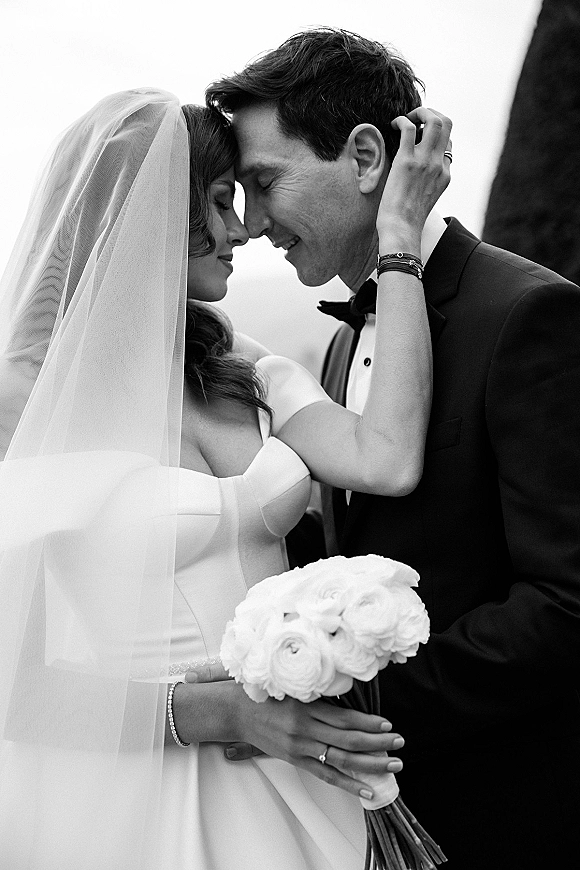 Wedding couple portrait in black and white, bride and groom touching foreheads as she holds a white bouquet, veil against blurred sky