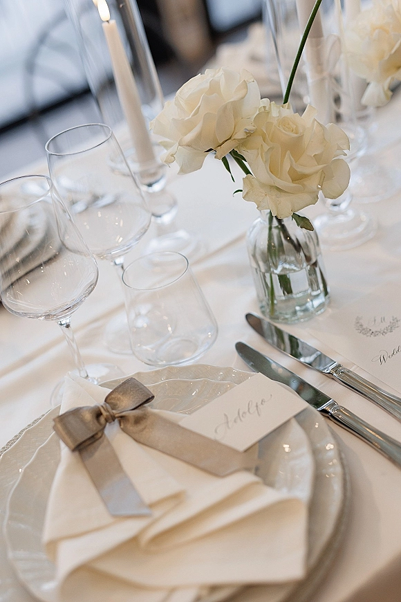 Reception tablescape with wedding place setting, white roses in bud vases, taper candles, and calligraphy place cards in an indoor space