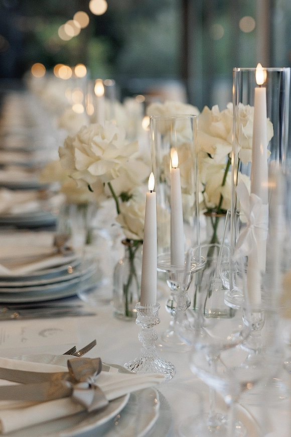 Reception tablescape with a white wedding centerpiece, taper candles in glass hurricanes, crystal candlesticks, and ribbon-tied napkins under string lights