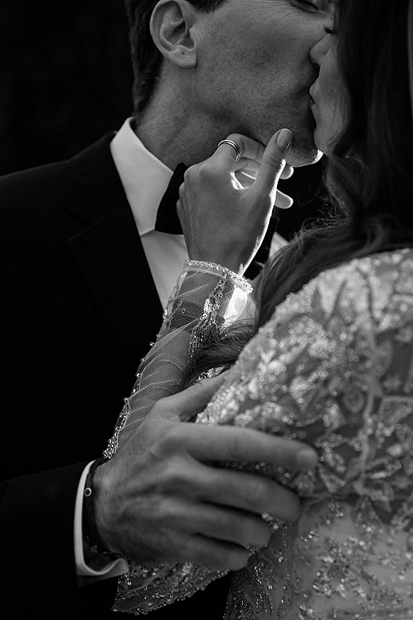 Wedding kiss portrait in close up as bride and groom kiss, her ringed hand on his face, tuxedo bow tie and beaded gown against dark blur