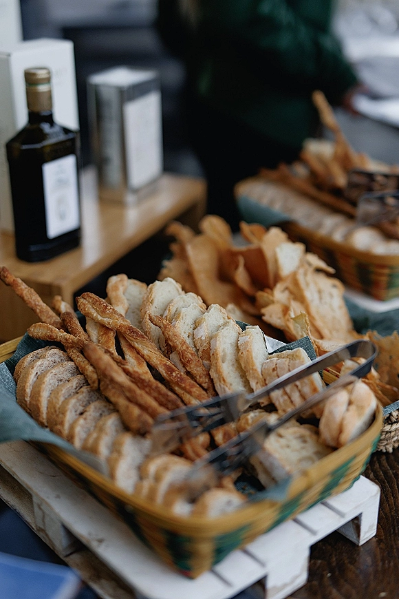 Wedding bread display with wedding bread basket assortment in wicker trays, napkins and tongs on a wooden table in a blurred indoor catering area