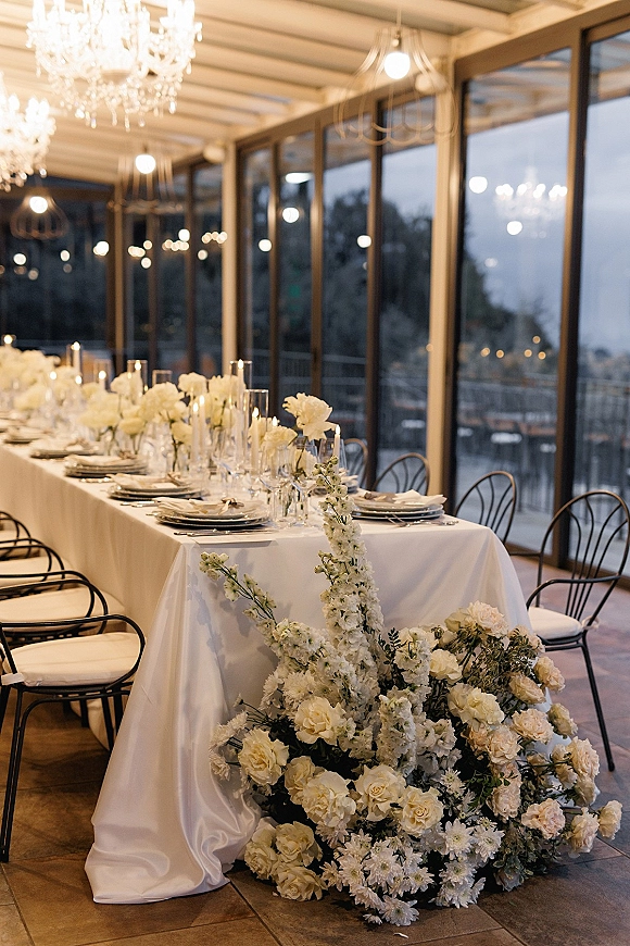 Reception tablescape with head table decor featuring all-white floral arrangements, taper candles, and place settings beneath chandeliers by glass windows