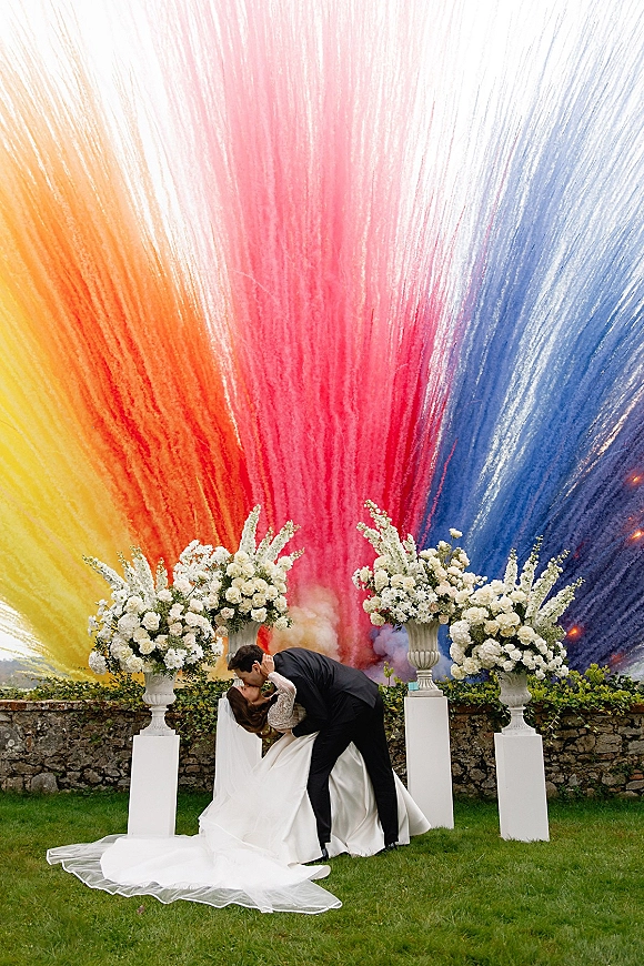 Wedding kiss as the groom dips the bride in a cathedral veil, with white urn florals and a rainbow smoke cloud on a garden lawn