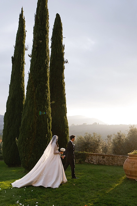 Couple portrait of bride and groom walking hand in hand, her veil trailing behind, at sunset among cypress trees and a stone wall