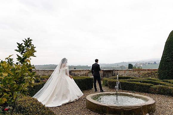 First look moment as bride walks up behind groom, her long veil trailing on a gravel garden path near a stone fountain and hedges
