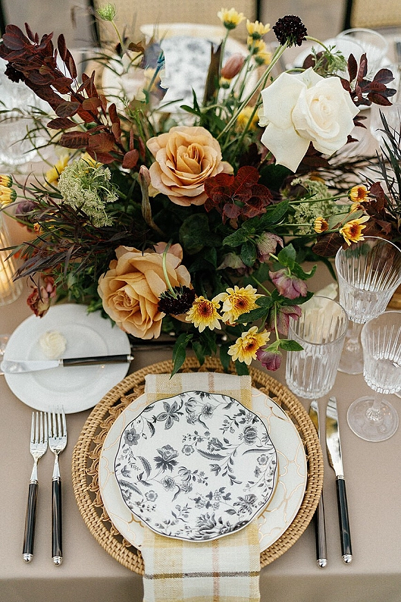 Reception tablescape with wedding table setting featuring rose and greenery floral centerpiece, patterned plates, woven chargers, and crystal goblets